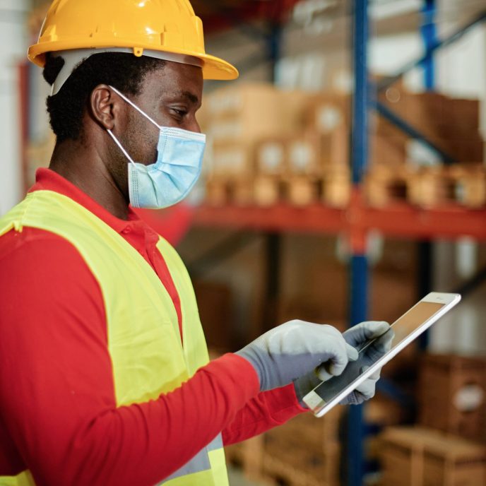 Caucasian adult warehouse worker checks delivery order on a tablet while wearing a safety mask - Focus on the Face